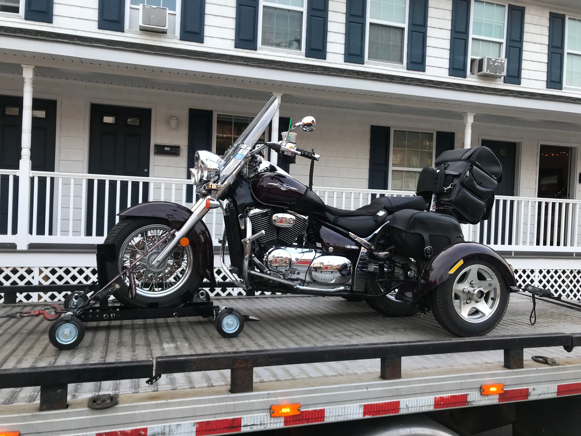 Black trike motorcycle on a tow truck, parked in front of a white house with blue shutters.