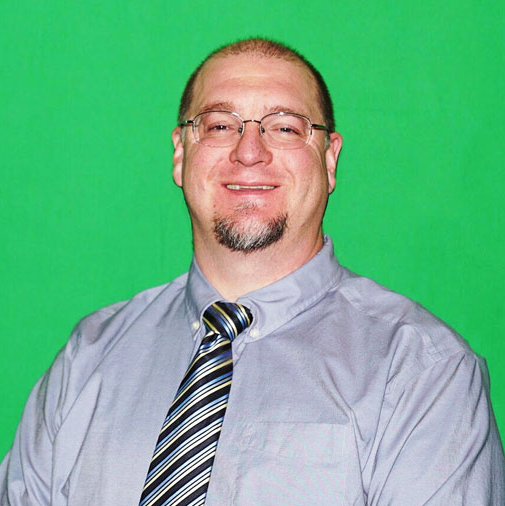 Man with glasses, goatee, and tie smiling in front of a green screen.