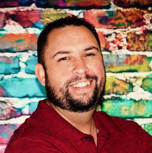 Man with a beard smiles, wearing a red shirt, in front of a colorful brick wall.