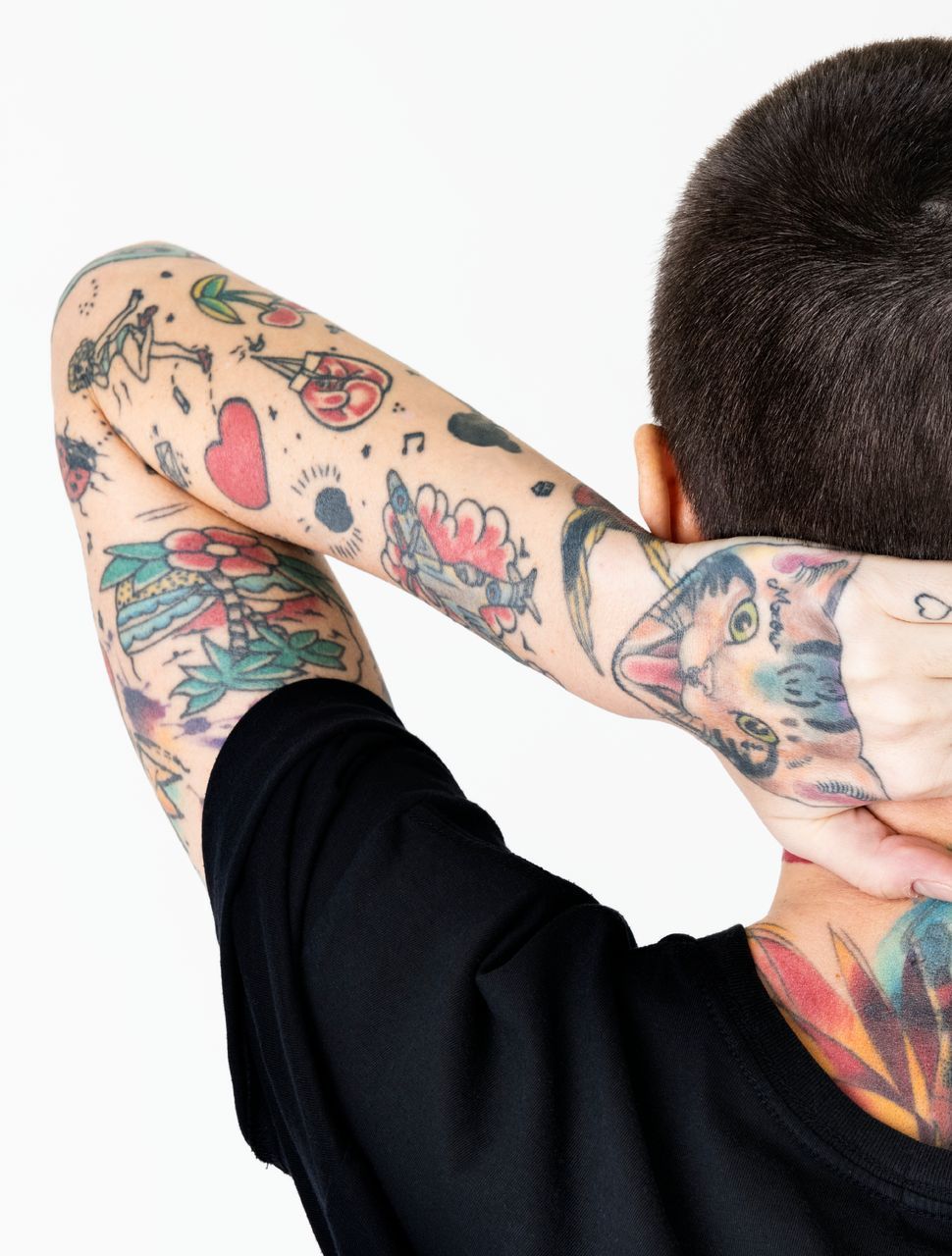 Person with arm tattoos, black shirt, hands behind head, neck tattoo, white background.