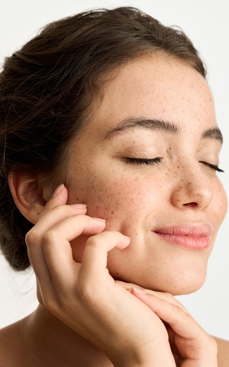 Woman with freckles smiles, eyes closed, touching her cheek.