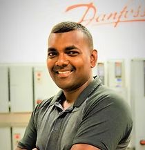 A man in a grey shirt is standing with his arms crossed and smiling.