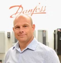 A bald man in a blue shirt is standing in front of a danfoss sign.