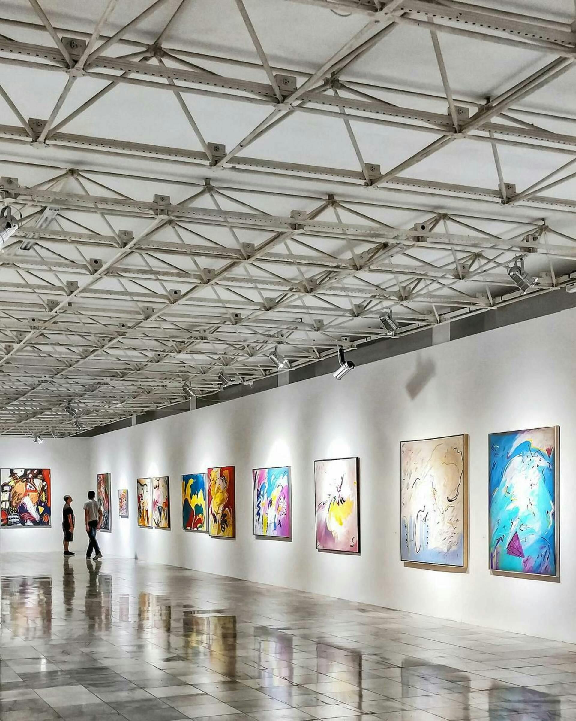 Art gallery with paintings on white walls, geometric ceiling, people viewing.