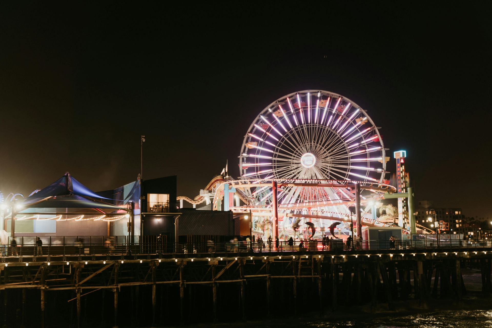 Ferris wheel lit up at night on a pier, with other lit buildings and silhouettes of people.