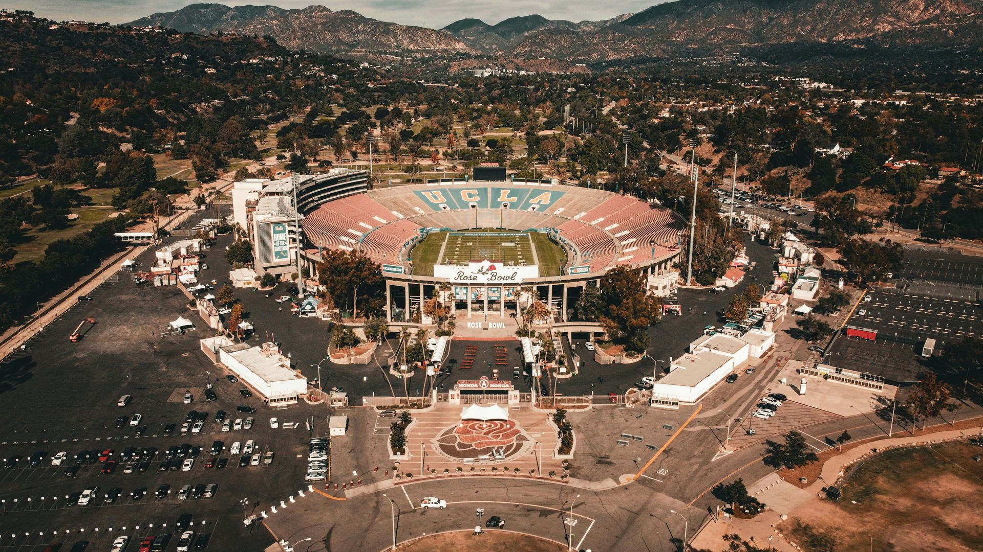 Aerial view of the Rose Bowl stadium in Pasadena, California. Green football field, red and beige seating, parking, and surrounding hills.