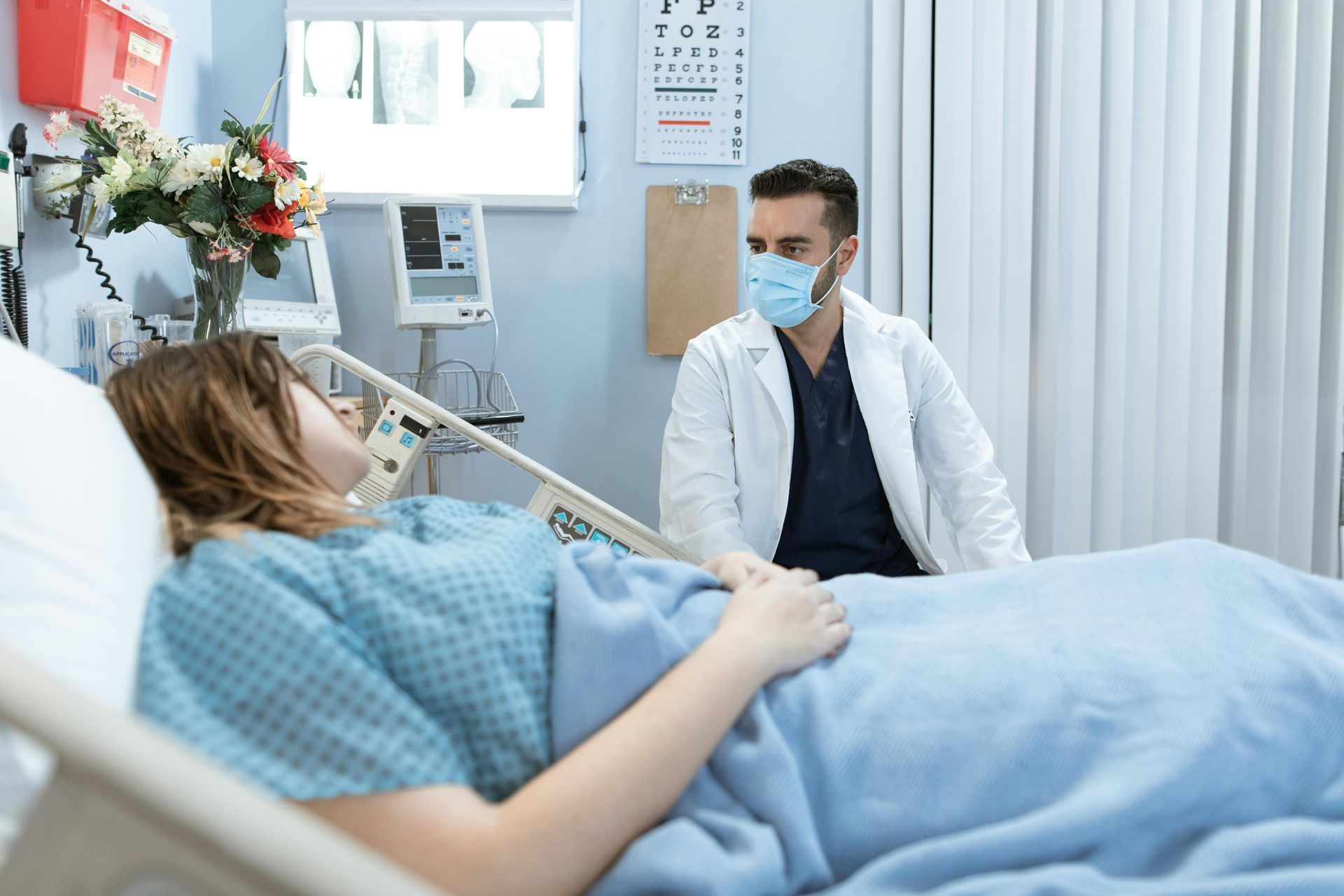 Doctor in mask talks to patient in hospital bed.