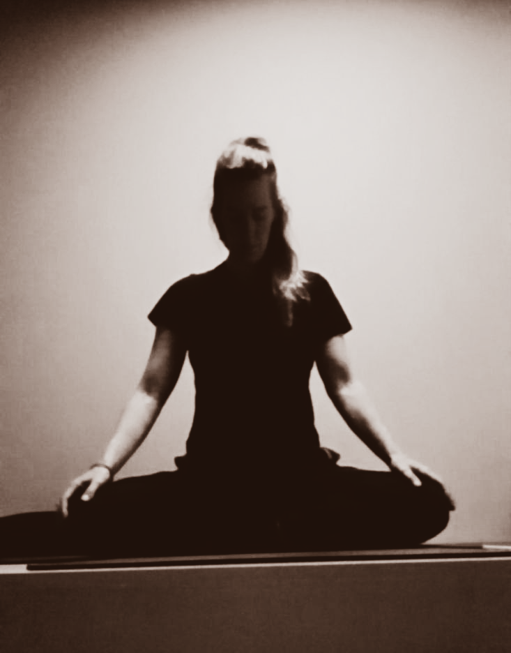 Vrouw in yogahouding, ogen gesloten, handen rustend op de knieƫn, zittend met gekruiste benen, zachte verlichting.