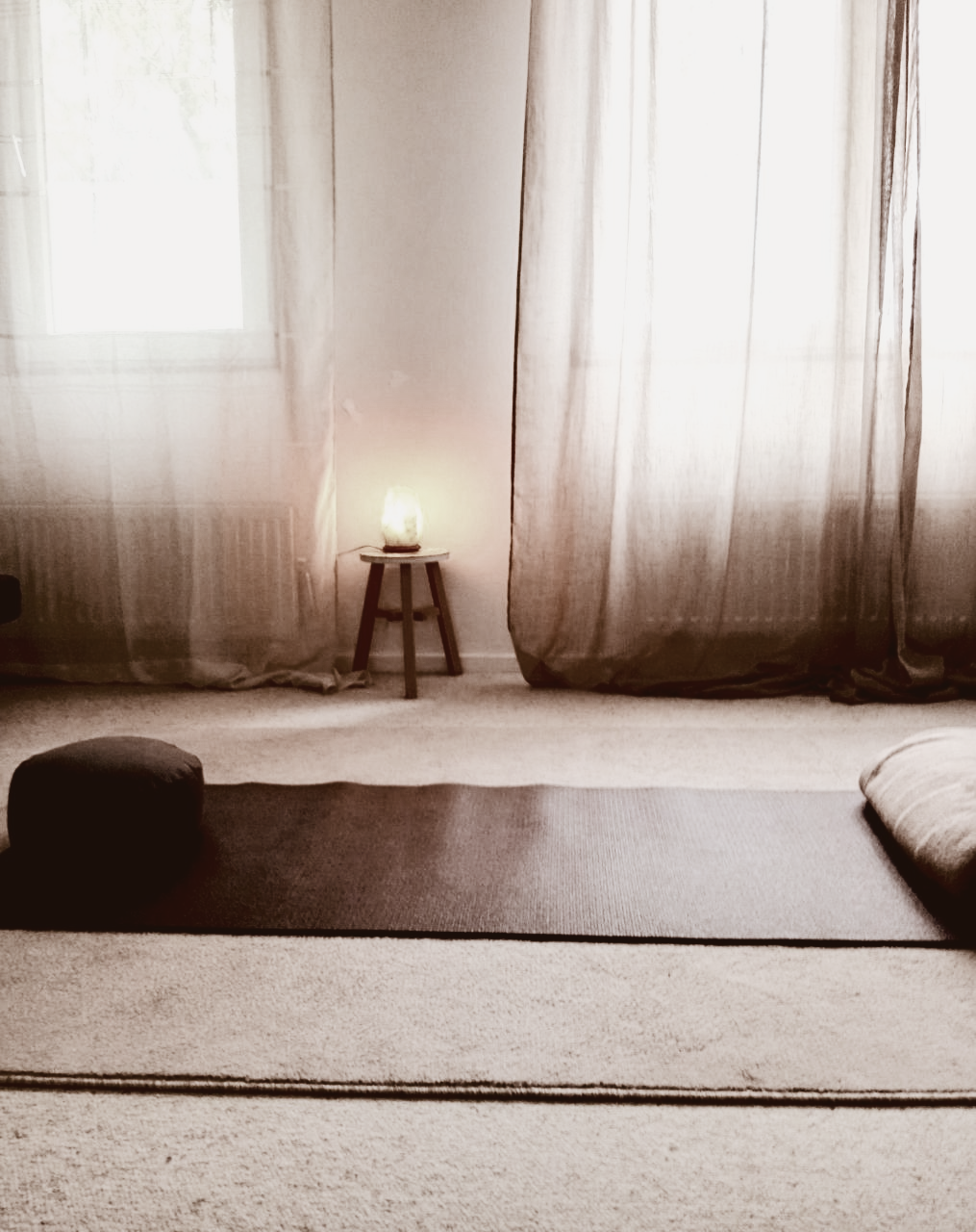 Een yogamat en -kussen in een lichte kamer met doorschijnende gordijnen en een klein tafeltje met een lamp.