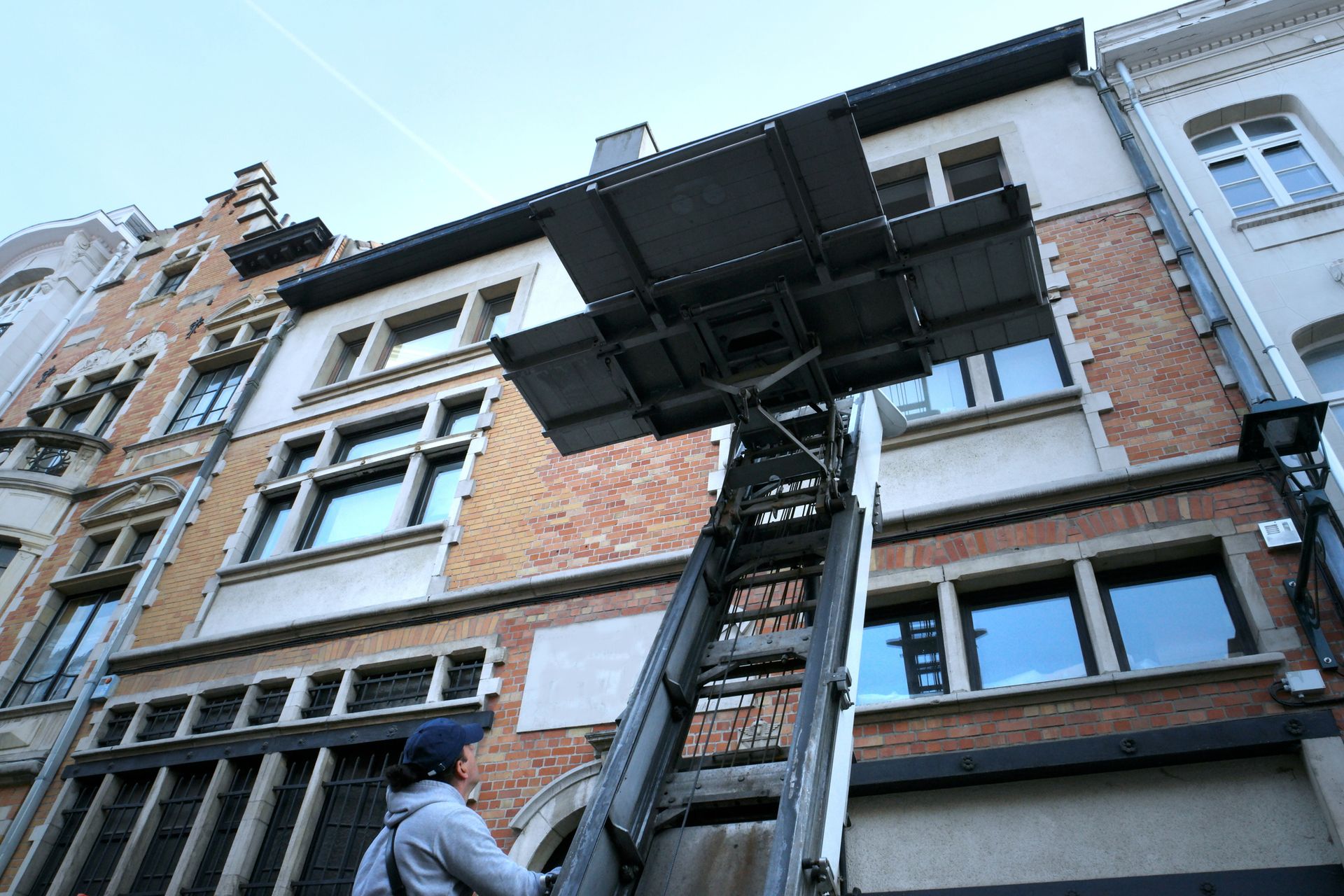 Una persona aziona una piattaforma elevatrice per spostare oggetti in un edificio con finestre e dettagli architettonici.