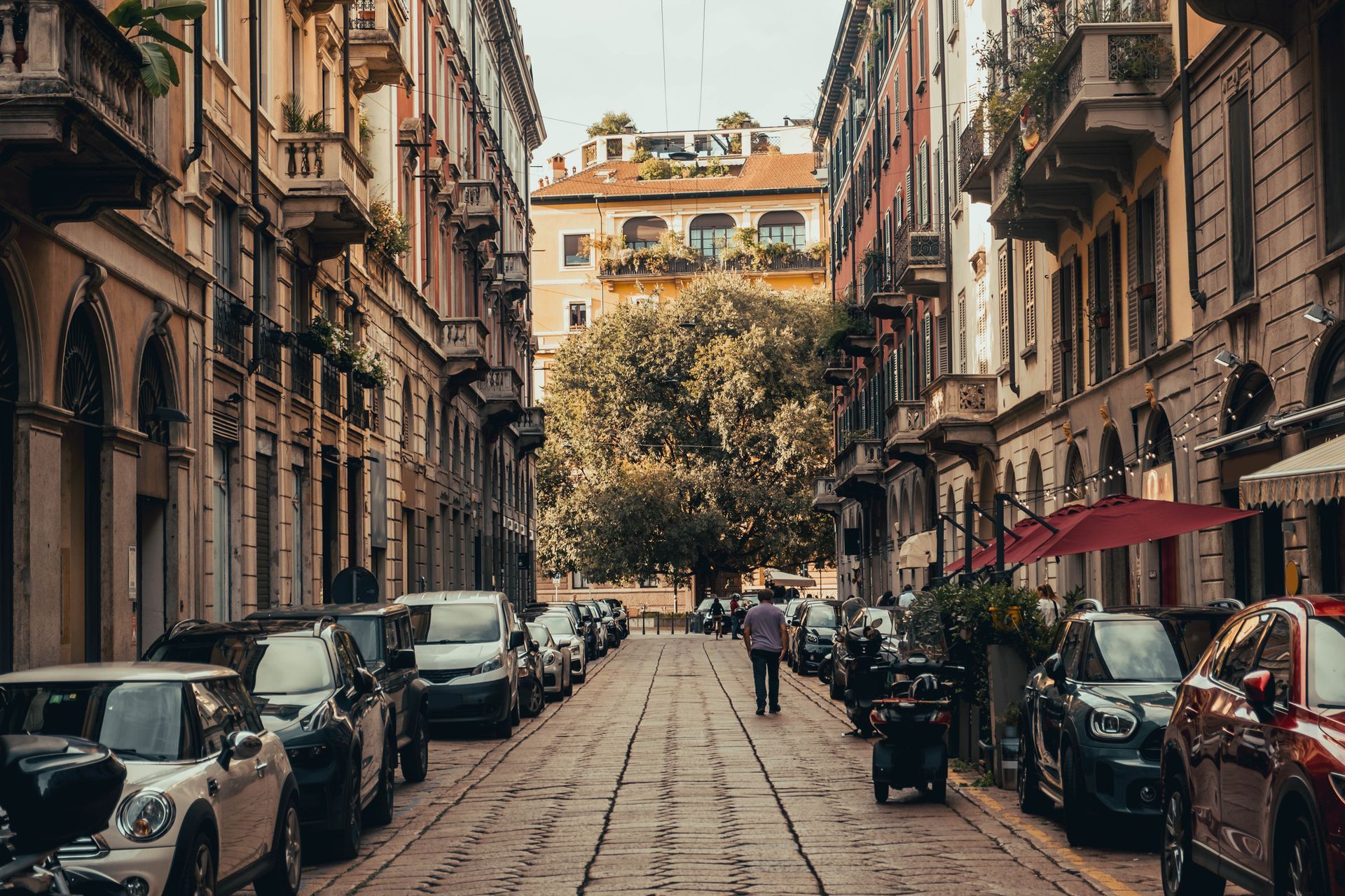 Strada stretta a Milano, Italia, fiancheggiata da edifici e auto parcheggiate; una persona cammina verso un albero in fondo.