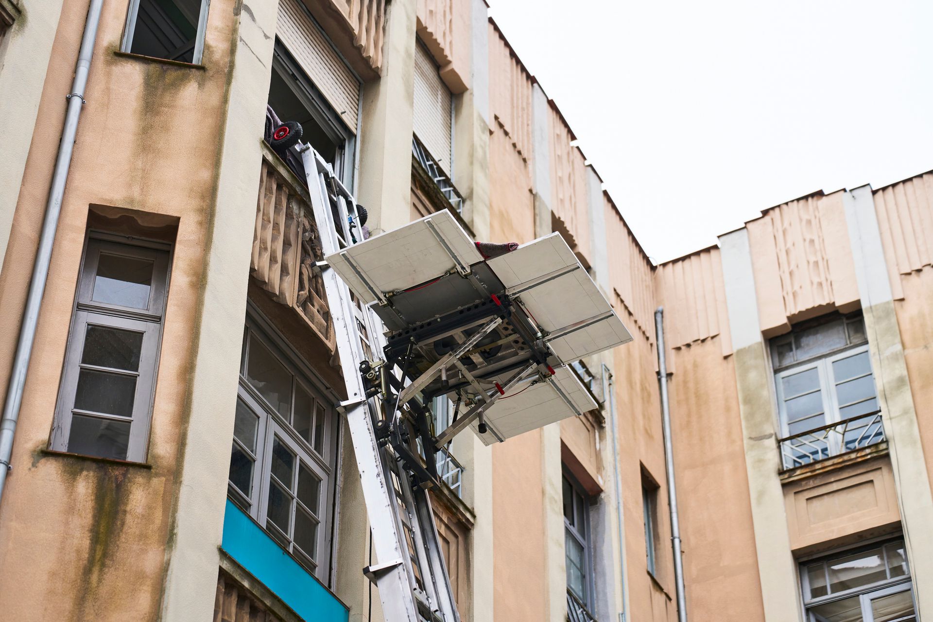 Una piattaforma elevatrice si estende fino alla finestra di un appartamento. Esterno dell'edificio beige con numerose finestre.