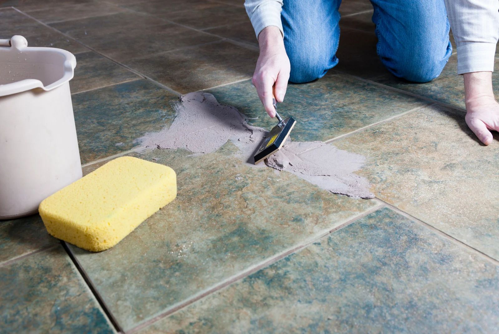 Person kneeling, applying grout to tile floor with a trowel, next to a bucket and sponge.
