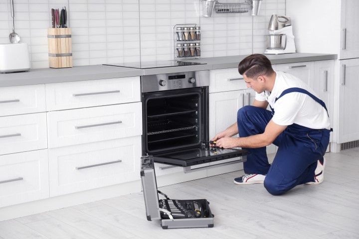 A technician in blue work overalls kneels on a kitchen floor, repairing an open oven with a nearby metal tool kit.