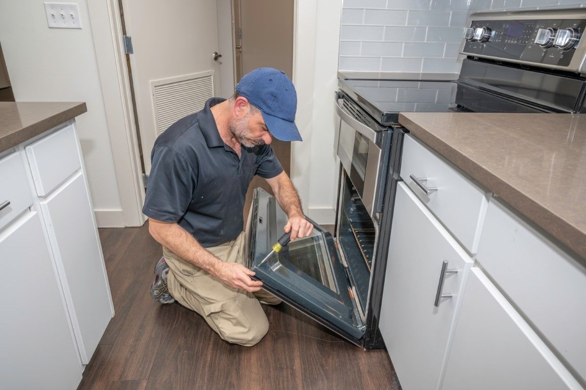 A technician in a blue cap kneeling on a kitchen floor while repairing the open door of a modern oven.