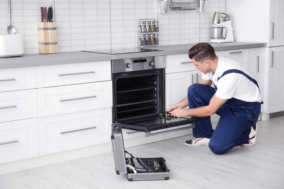 A technician in blue overalls kneeling on a kitchen floor while repairing an open oven with a tool case nearby.