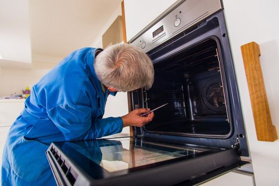 A person in a blue uniform inspects the interior of a wall-mounted oven.