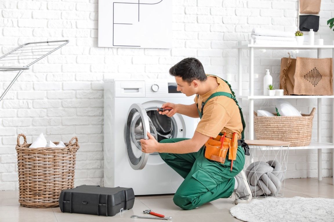 A repair person in green coveralls kneeling to fix a white washing machine in a bright laundry room.