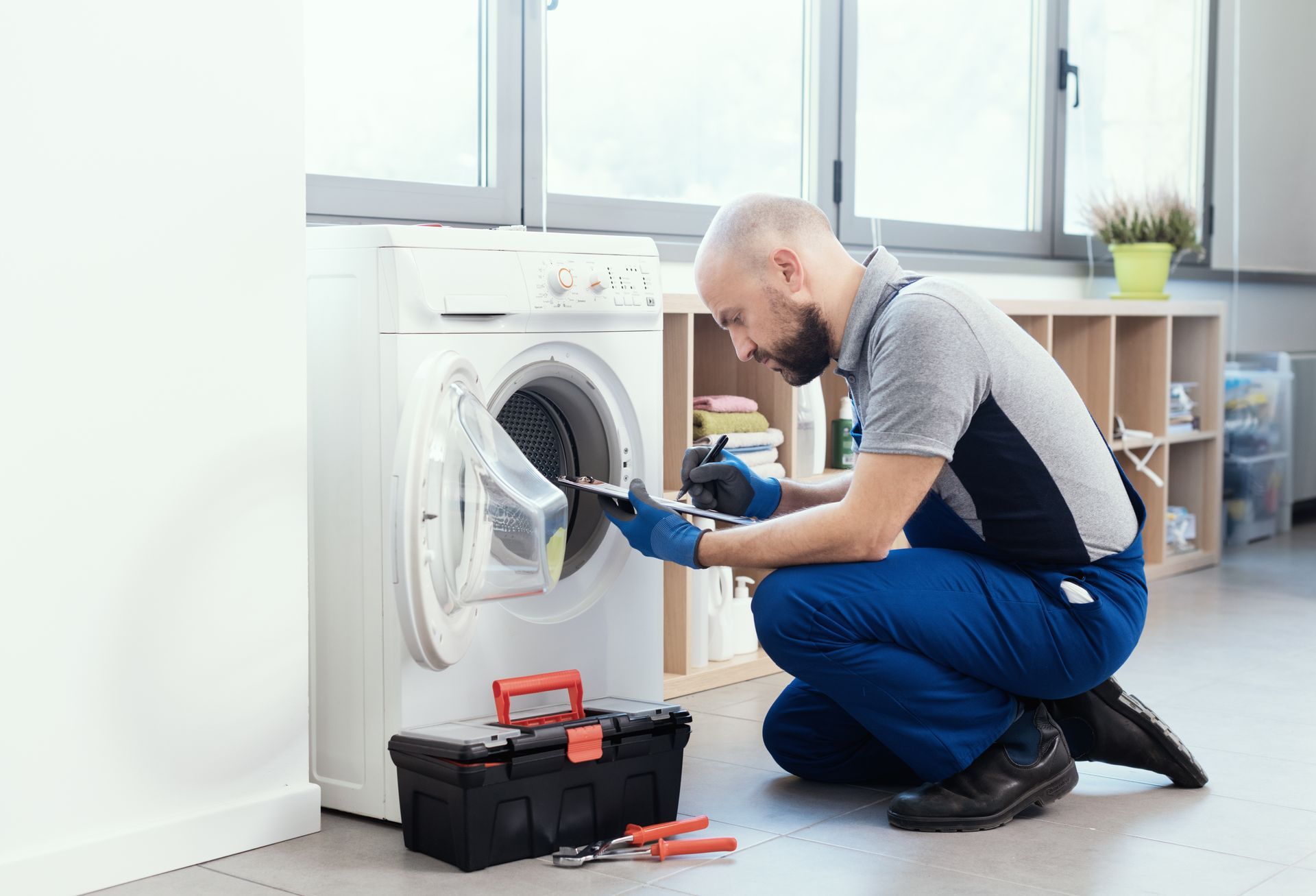 A technician in blue overalls kneels on the floor, using a tool to repair the door of a white washing machine.