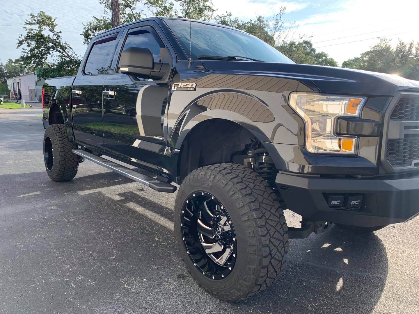Black Ford F-150 pickup truck with large tires, parked on pavement. Sunny day.