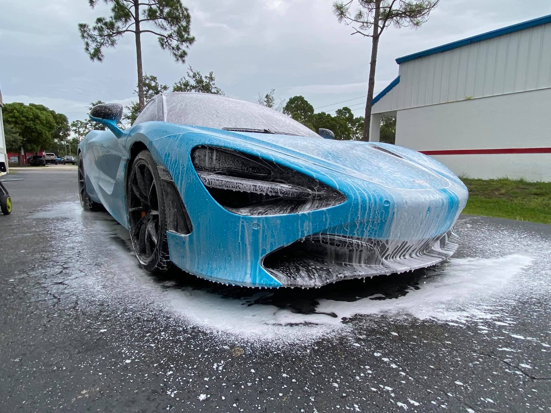 A teal McLaren sports car covered in white foam, being washed outdoors.
