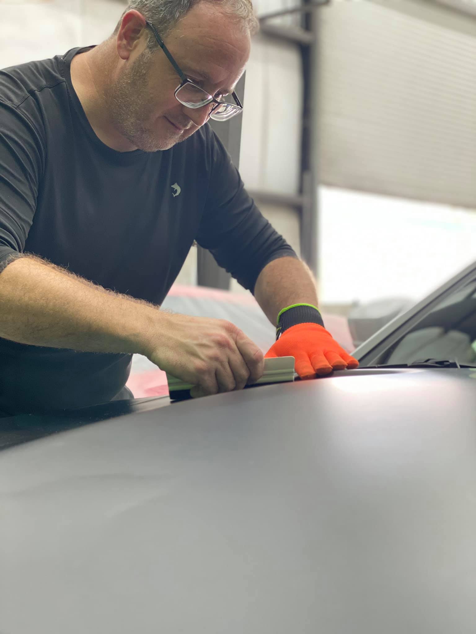 Man with glasses wearing orange gloves working on a car's hood.