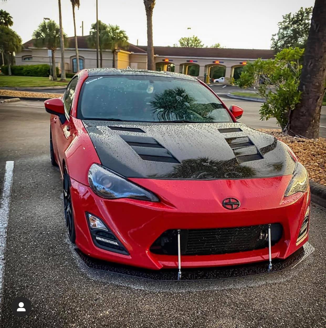 Red Scion FR-S sports car with a black carbon fiber hood, parked in a lot with palm trees.