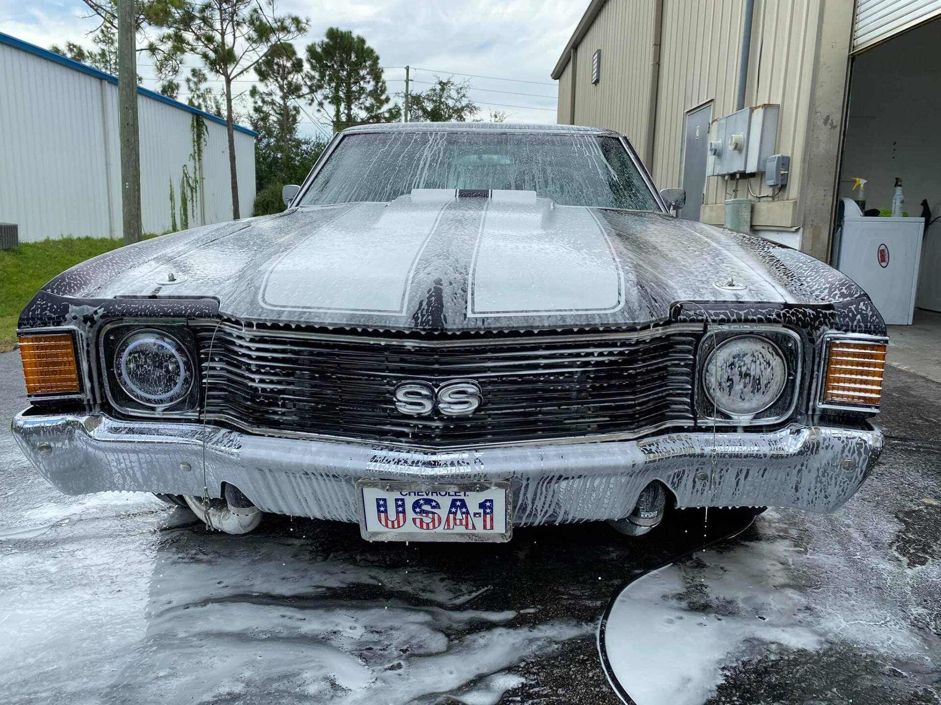 Black classic car, covered in white foam, being washed outdoors.
