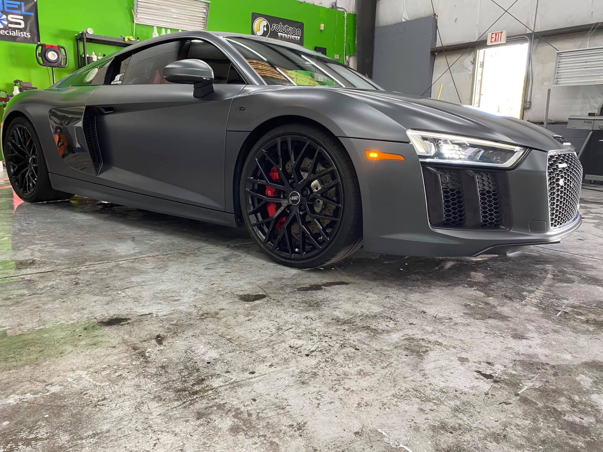 Dark gray Audi R8 sports car, black wheels, red brake calipers, inside a shop.