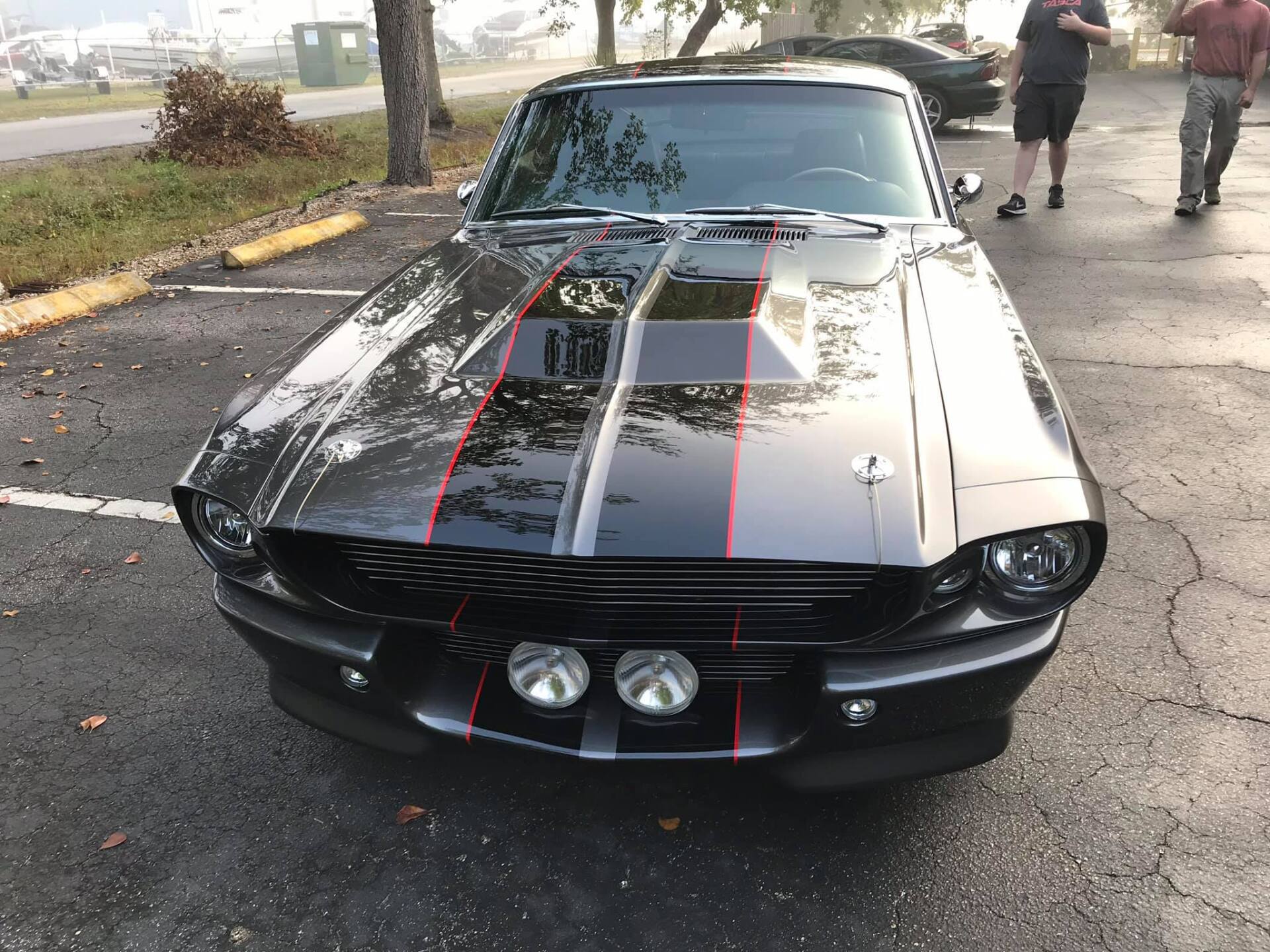 Black and gray vintage Ford Mustang with red stripes, parked, with two people walking in the background.