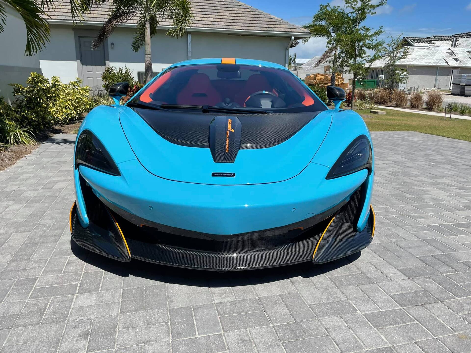 Blue McLaren sports car parked on a brick driveway. It has orange accents and a black roof.