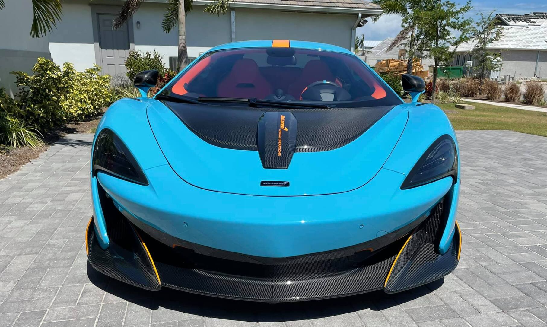 Blue McLaren sports car with orange accents parked in front of a house on a sunny day.