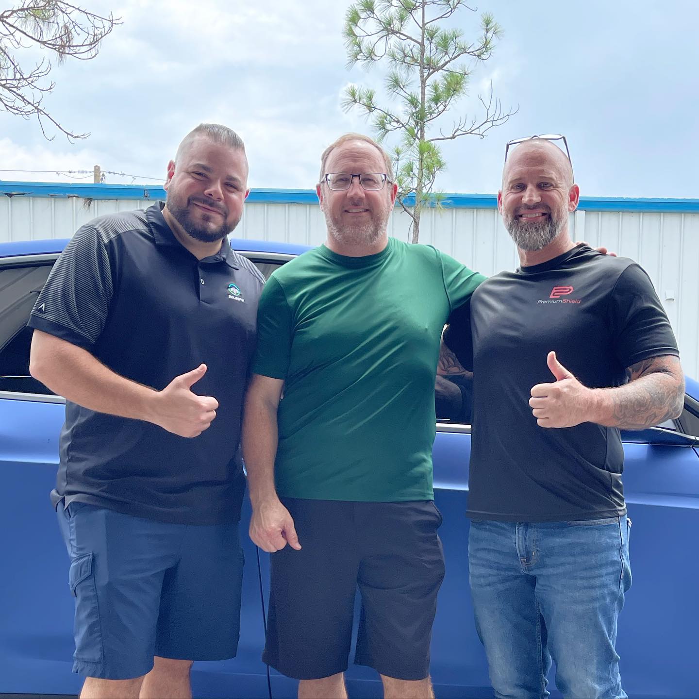 Three men, thumbs up, smiling in front of a blue car. Sunny outdoor setting.