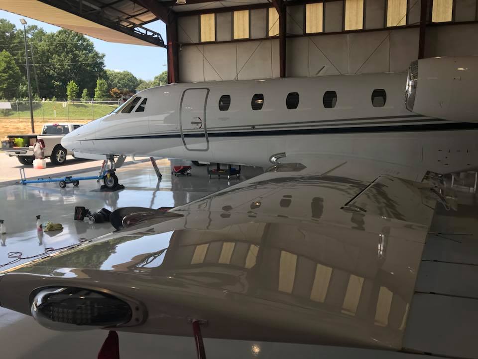 A white private jet inside a hangar, the wing in foreground reflecting the aircraft and its surroundings.
