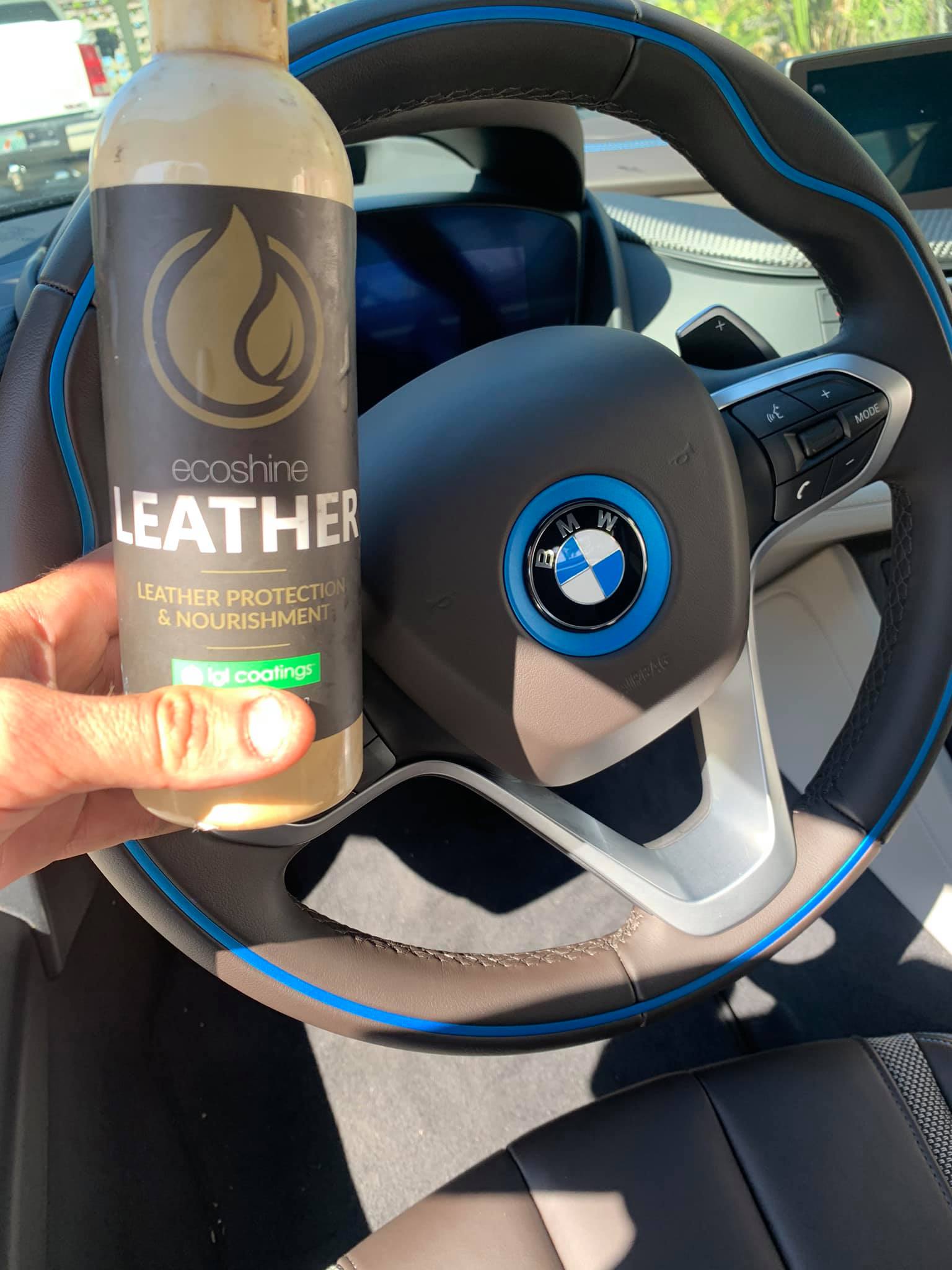 A hand holds a leather protectant bottle in front of a BMW steering wheel. The bottle has a brown label and logo.