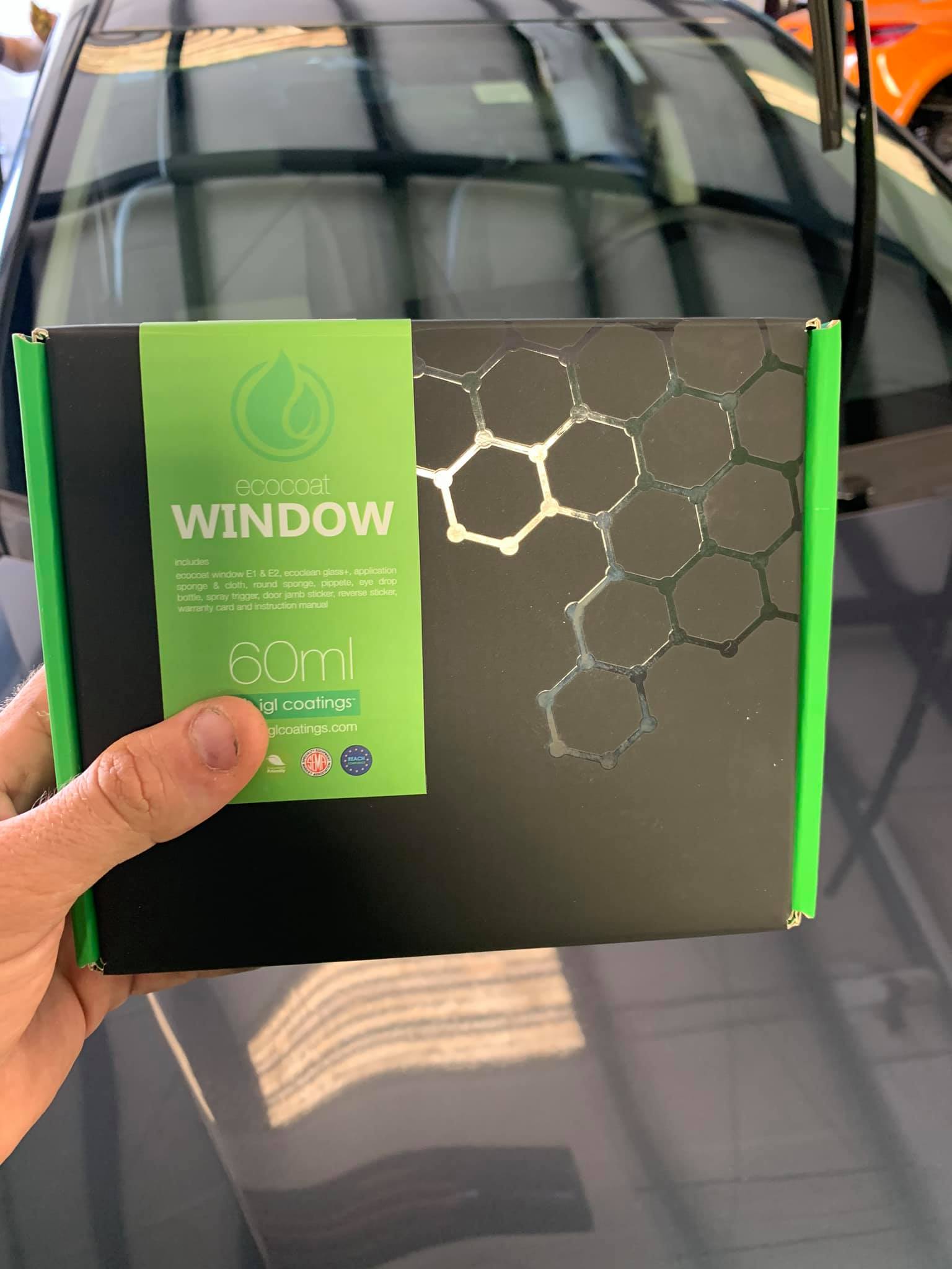 Hand holding a box of window cleaning product with a car in the background. The box is black with green accents.