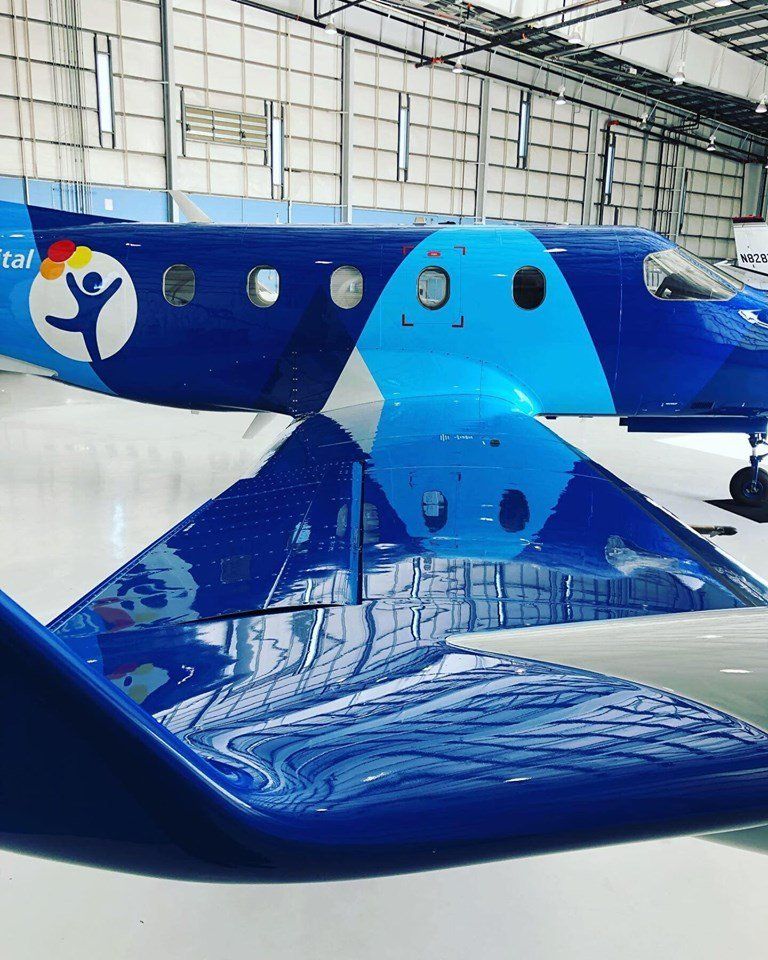 Blue and white airplane with a charity logo, reflected on a shiny surface in a hangar.