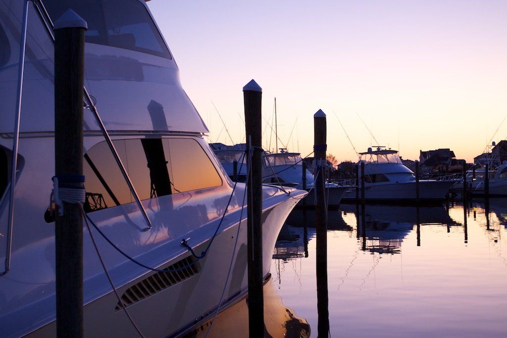 A white yacht docked in a harbor at sunset; purple and orange hues reflect in the water.