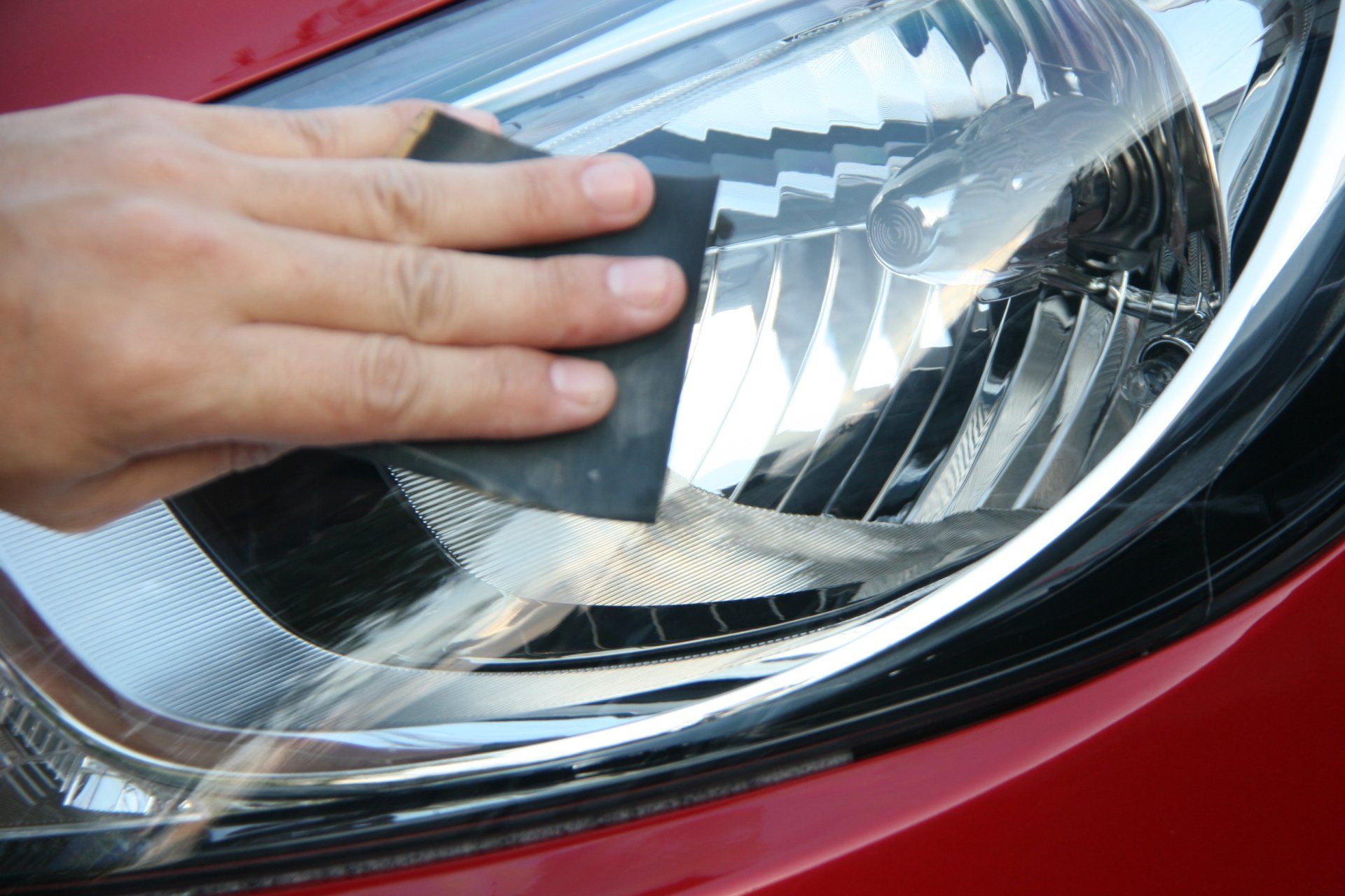 Person sanding a cloudy car headlight with a black abrasive pad; red car in daylight.