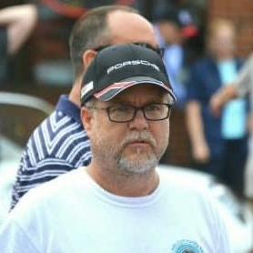 Man wearing glasses and a Porsche cap smiles, with a second man in the background.