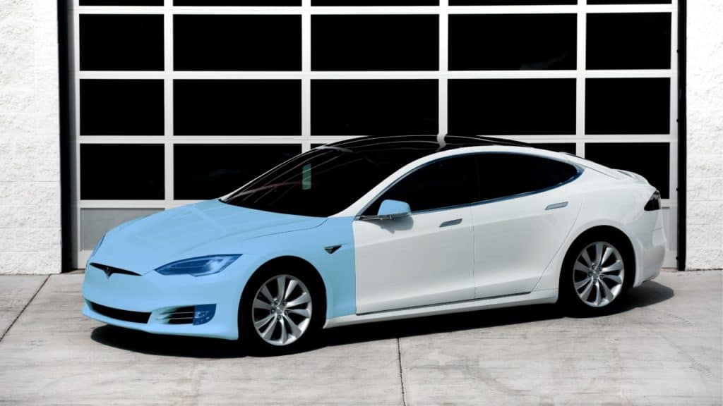 White and light blue Tesla car parked in front of a garage with a black and white door.