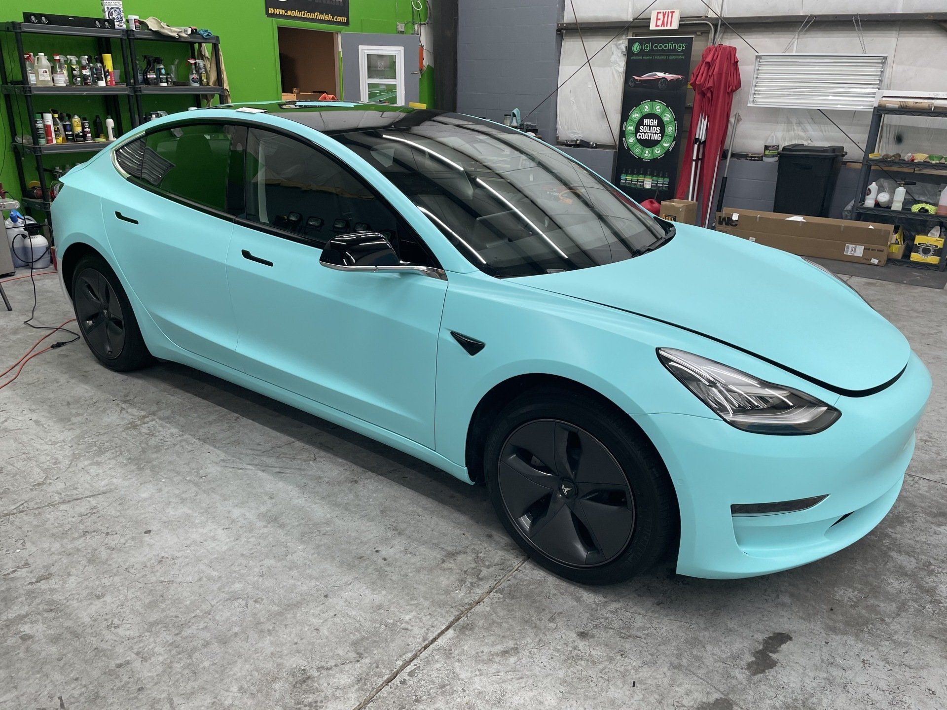 A teal Tesla Model 3 car in a garage. It has a black roof and black wheels.