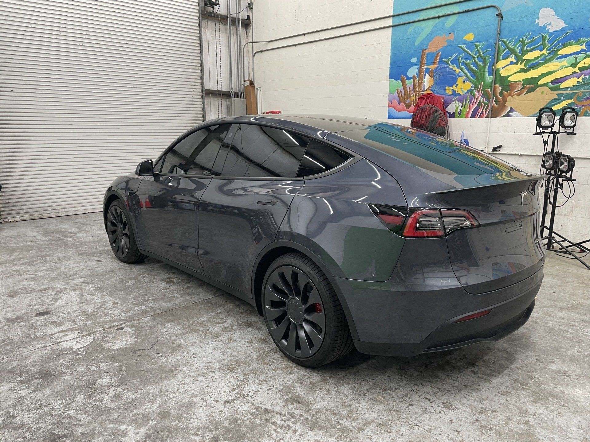 Dark gray Tesla Model Y in a garage, angled shot, tinted windows, black wheels.