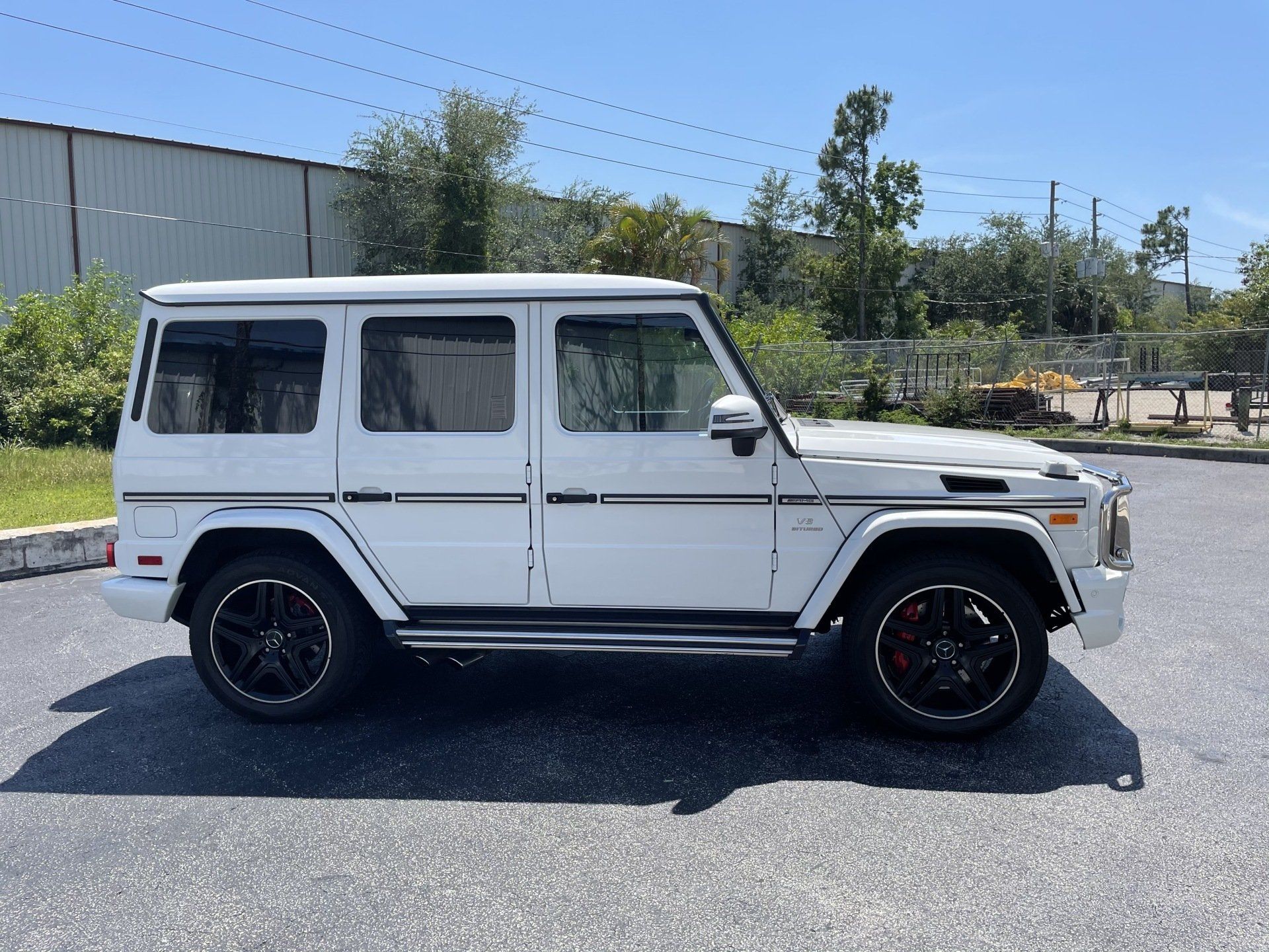 White Mercedes-Benz G-Class SUV parked on asphalt with black rims and a commercial building in the background.