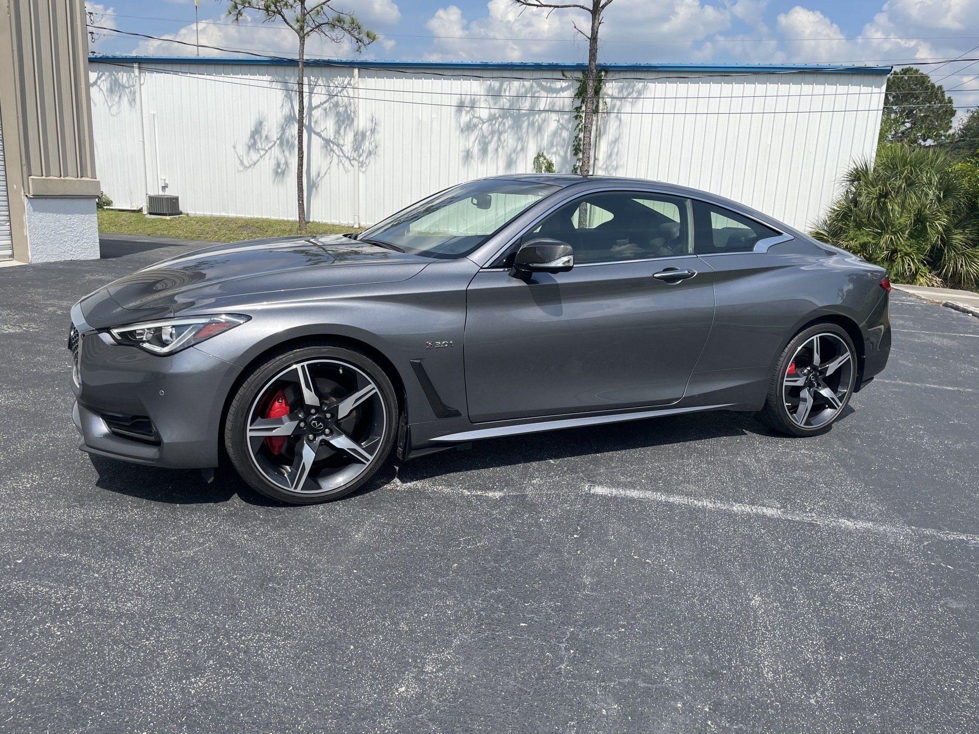 Gray Infiniti Q60 coupe parked on asphalt, red brake calipers, in front of a white building.