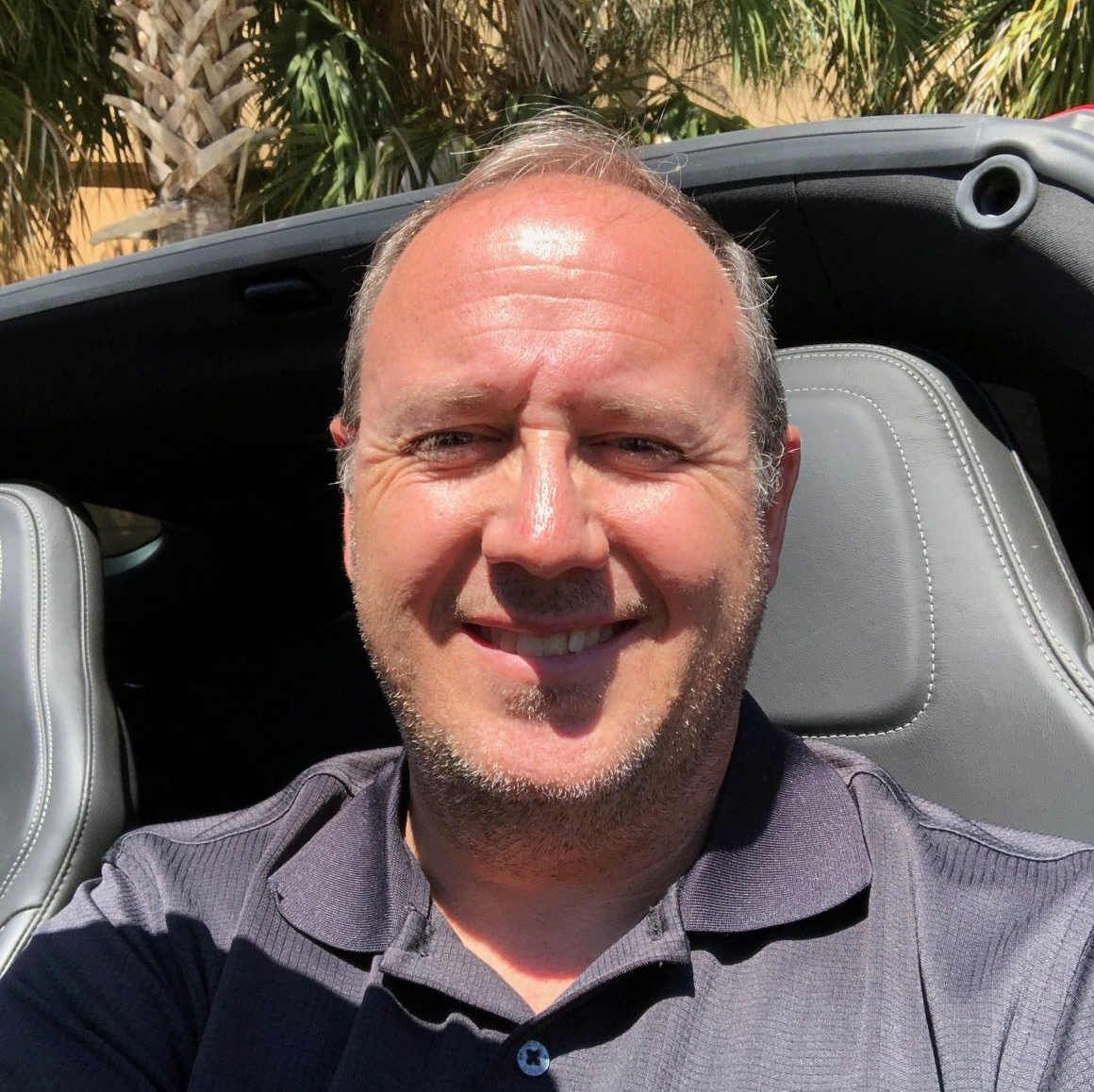 Man smiling, sitting in a car, wearing a gray polo shirt, outdoors.