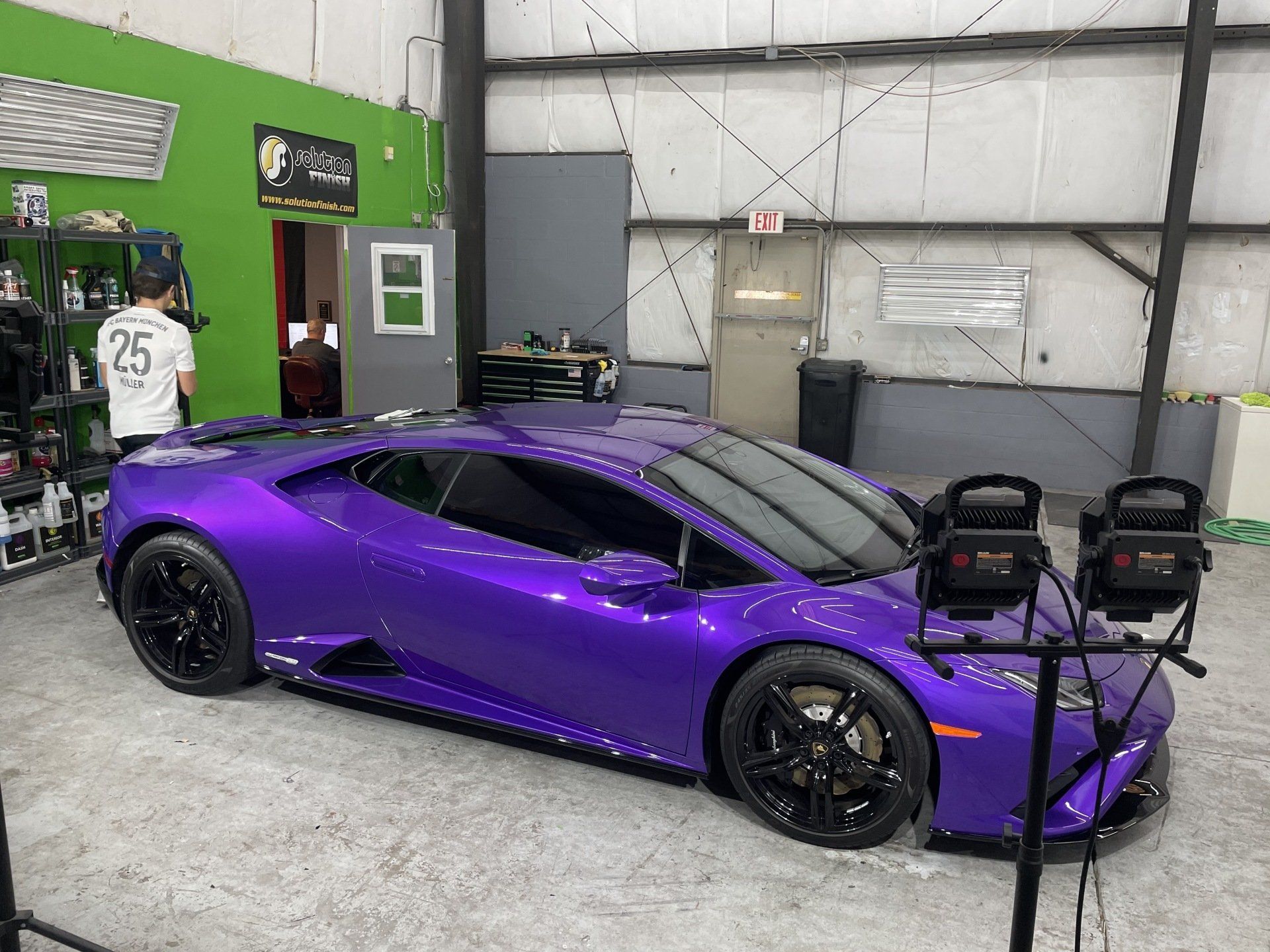 Purple Lamborghini in a garage, illuminated by lights, with a person in the background.