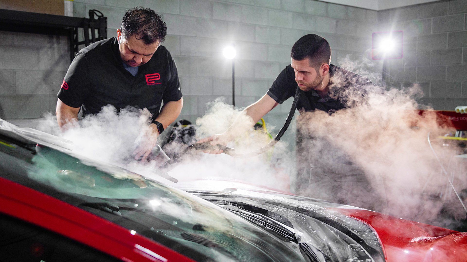 Two men steam cleaning a red car in a garage.