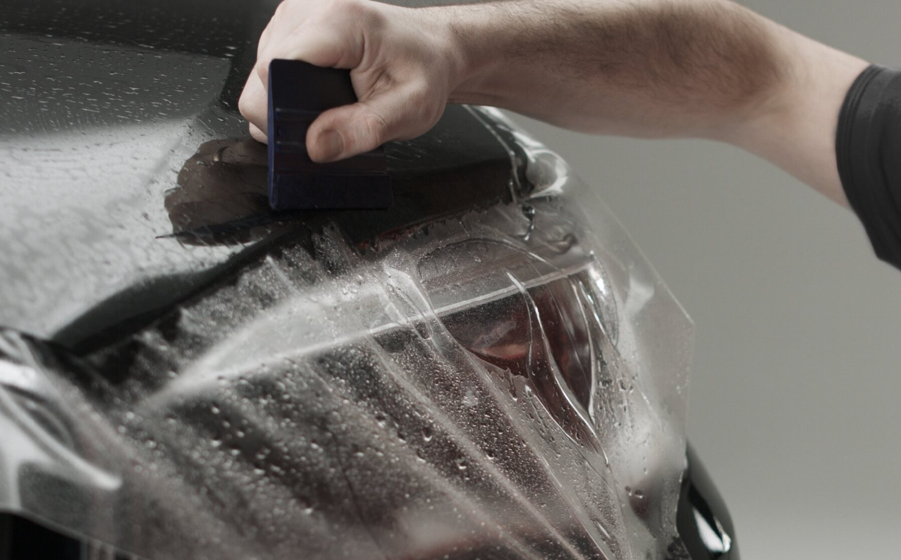 Person applying film to a car headlight with a squeegee; the setting is a studio.