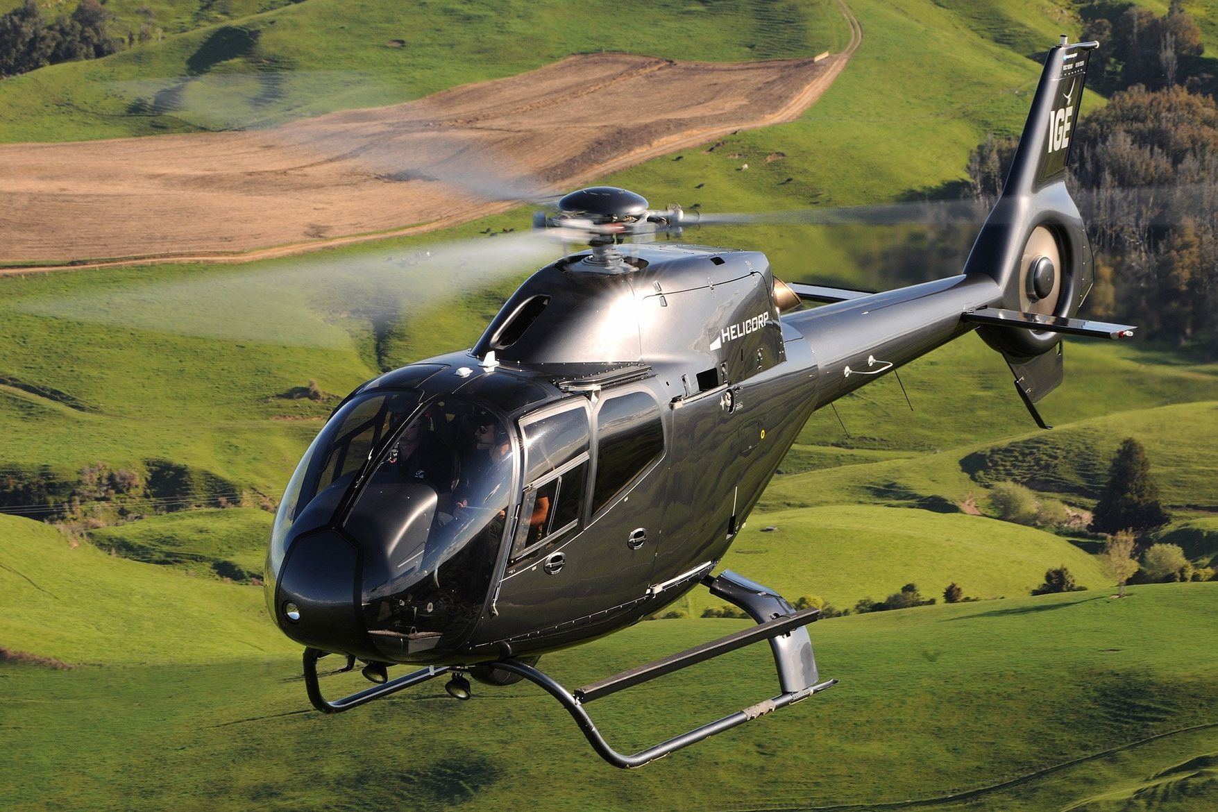 Black helicopter flying over green hills.